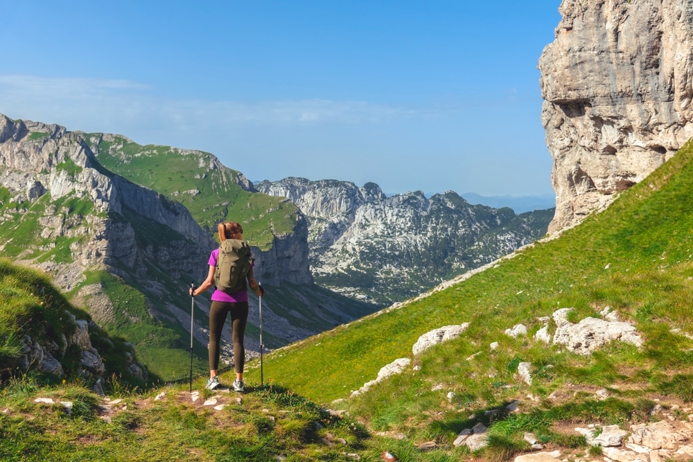 Back view of young female traveler with trekking sticks hiking in Alps.