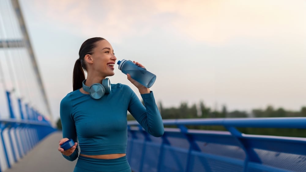 Fit European woman relaxing and drinking water on seaside promenade after running and training