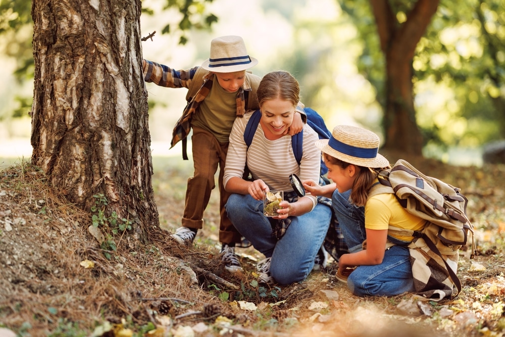 two kids boy and girl with backpacks looking examining environment through magnifying glass while exploring forest nature on sunny day